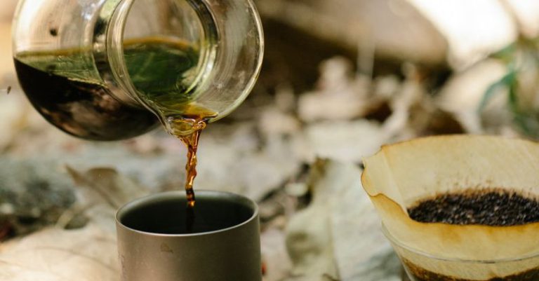 Morning Hikes - Crop unrecognizable male hiker pouring fresh hot coffee from chemex into metal mug placed on ground covered with dry fallen leaves at campsite