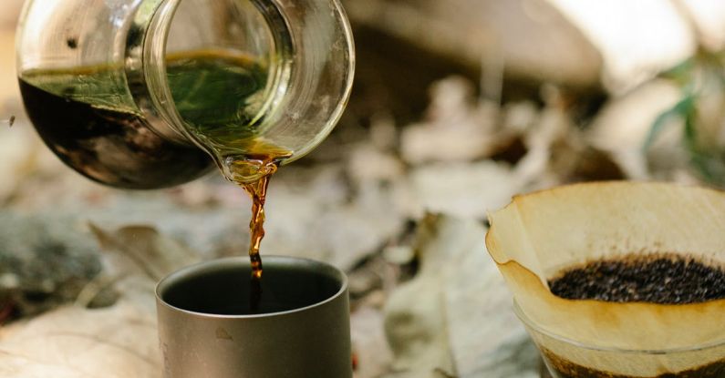 Morning Hikes - Crop unrecognizable male hiker pouring fresh hot coffee from chemex into metal mug placed on ground covered with dry fallen leaves at campsite