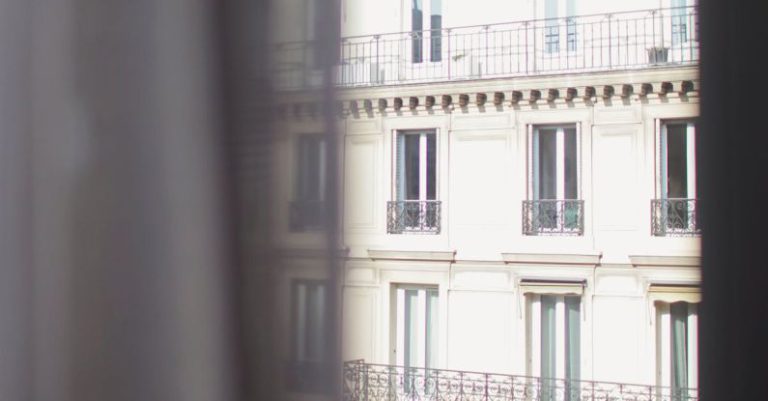 Hotel Stay - Hotel with elegant balconies and bay windows in Paris as seen from opposite building through slightly ajar window curtains on sunny day