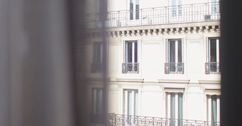 Hotel Stay - Hotel with elegant balconies and bay windows in Paris as seen from opposite building through slightly ajar window curtains on sunny day