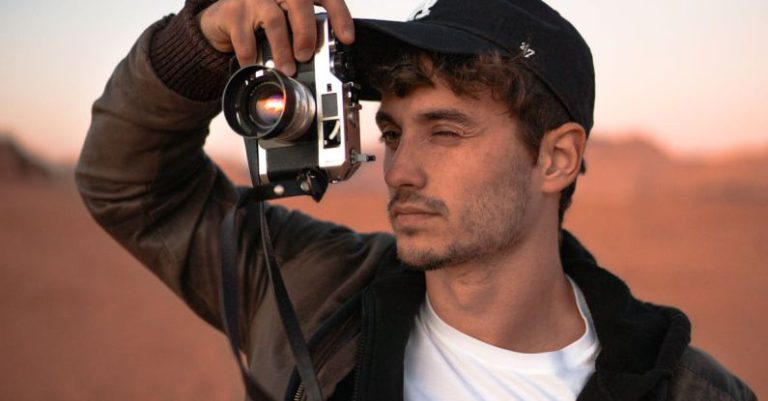 Successful Day Trip - Focused young male traveler in stylish outfit and cap taking photo on vintage camera during trip in desert valley during sunset